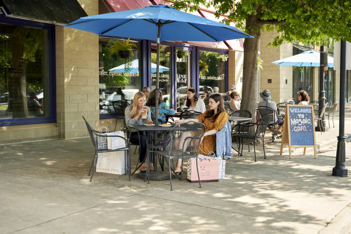Women sit outside Mosaic Cafe under a blue umbrella with food and shopping bags.